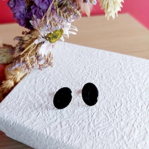 Op de afbeelding: Twee glanzende zwarte ovale oorbellen liggen op een wit, getextureerd oppervlak. De oorbellen staan centraal, met gedroogde bloemen in paars, wit en beige op de achtergrond.