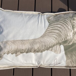 May include: Rectangular decorative pillow with a detailed illustration of an elephant's trunk and head in shades of gray and white. The pillow has a white background and a cream-colored border. The artist's signature is visible.