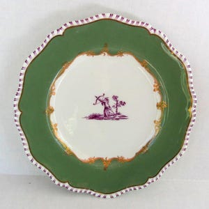 May include: A round ceramic plate with a scalloped edge featuring a wide sage green border and a white center. The rim has purple and white striped detailing. The center displays a purple illustration of an archer with gold scrollwork accents.