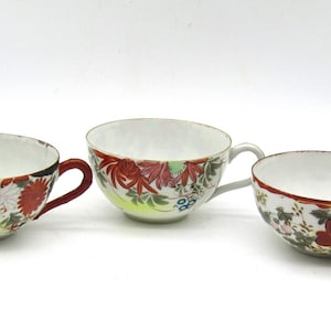 May include: Three white porcelain teacups with red handles and floral designs. The cups have a white background with red, pink, green, and blue floral patterns.