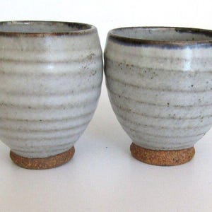 May include: Two ceramic cups with a light gray, speckled glaze and horizontal ribbed texture. Each cup features a dark, brownish-gray rim and a distinct, unglazed, textured brown base that contrasts with the smooth, lighter body of the vessels.