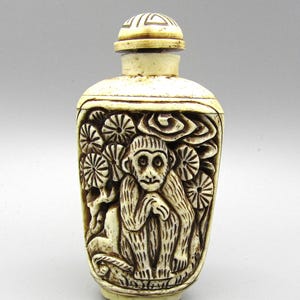 May include: An off-white, rectangular bottle with a detailed relief carving of a monkey surrounded by flowers and clouds. The bottle has a matching cap with a geometric design. The carving is in a darker shade, creating contrast.