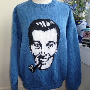 May include: A blue knitted sweater featuring a black and white portrait of a man with a pipe. The portrait is centered on the front of the sweater. The sweater has a crew neck and long sleeves.