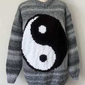 May include: A long-sleeve knit jumper featuring a large black and white yin-yang symbol centred on the front. The jumper is crafted from variegated yarn in shades of charcoal, slate grey, and light grey, with a ribbed crew neckline and cuffs.