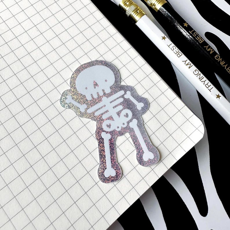May include: A holographic sticker of a stylized skeleton figure with a rounded head and simple bone structure. The sticker features a shimmering, multi-colored reflective surface and is placed on a white grid paper notebook page.