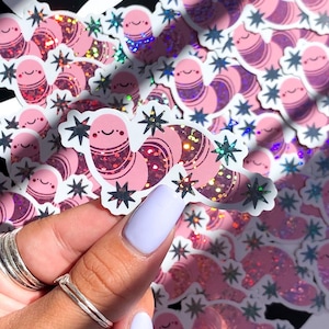 May include: A sheet of pink and purple glitter worm stickers with black star accents. The stickers are arranged in rows and are being held by a hand.