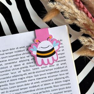 May include: A pink and black bee bookmark with a white face and yellow accents. The bookmark is on a page of a book with text.