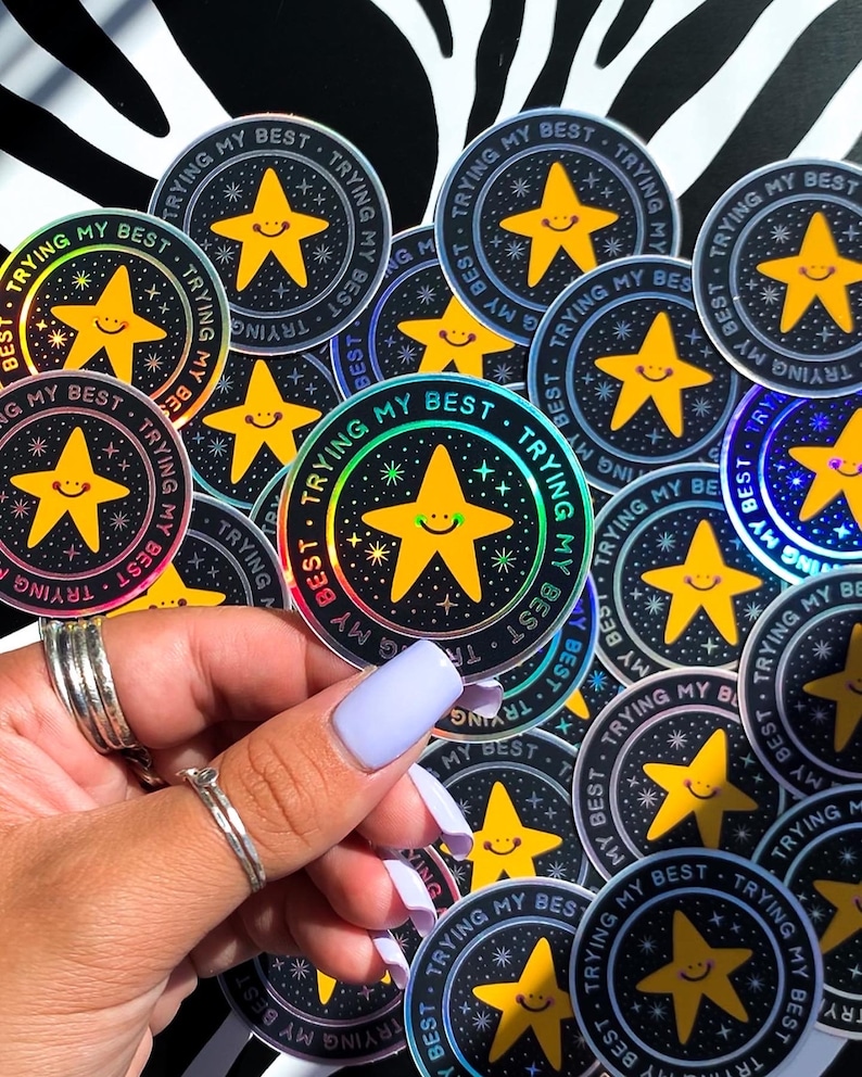 May include: A close-up of a hand holding a variety of iridescent stickers with a black background and a yellow star with a smiling face. The stickers say "Trying My Best" in a circle around the star.
