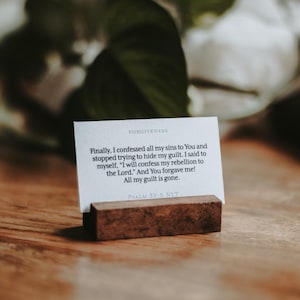 May include: A white card with text about forgiveness, held in a small, rectangular wooden block. The card is on a wooden surface, with greenery and white flowers in the background. The text on the card reads: "Finally, I confessed all my sins to You..."