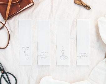 Puede incluir: Cuatro marcapáginas blancos con ilustraciones y texto en tinta negra. Los marcapáginas representan una mano sosteniendo un libro, una taza de café con vapor y el texto "one more page" y "cozy nights".