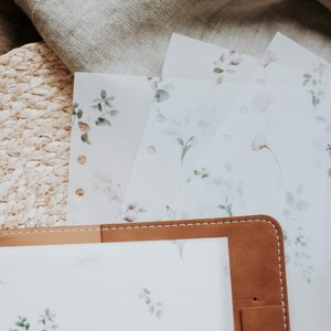 Puede incluir: Una agenda de cuero marr&oacute;n con una p&aacute;gina blanca con un patr&oacute;n floral. La agenda tiene una solapa que se cierra con un broche a presi&oacute;n. La agenda est&aacute; abierta para revelar una p&aacute;gina blanca con un patr&oacute;n floral.