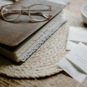 May include: A brown leather-bound journal with a clear plastic bookmark and a set of white index tabs. The journal is on a woven placemat with two white envelopes that say "Appoint" on them.