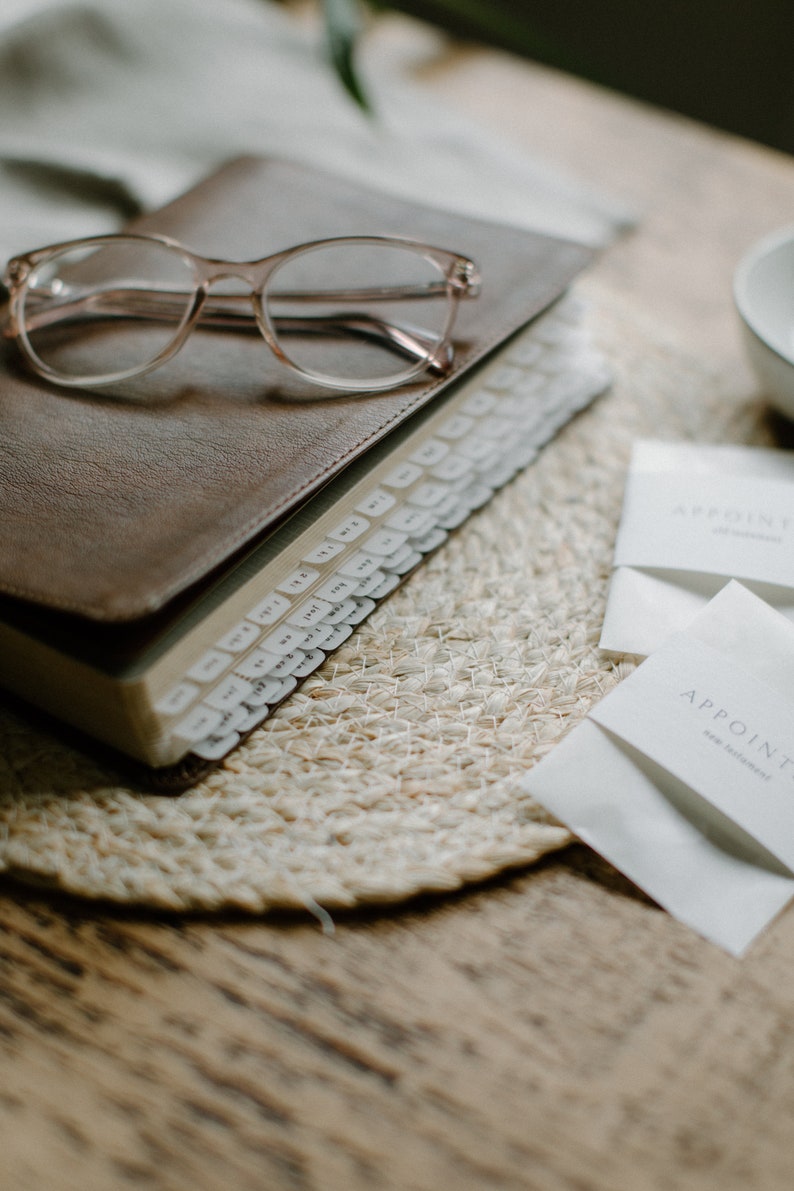 May include: A brown leather-bound journal with a clear plastic bookmark and a set of white index tabs. The journal is on a woven placemat with two white envelopes that say "Appoint" on them.