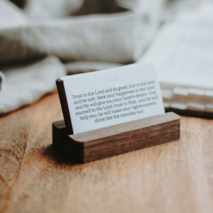 May include: A wooden card holder with a stack of white cards. The cards have text that reads "Trust in the Lord and do good; live in the land and be safe." The holder is made of dark wood.