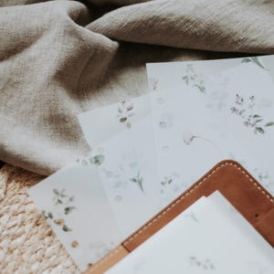 Puede incluir: Un diario de cuero marr&oacute;n con un inserto de papel blanco. El diario est&aacute; sobre una pila de hojas de papel blanco con un dise&ntilde;o floral. Las hojas est&aacute;n sobre una estera de paja tejida.