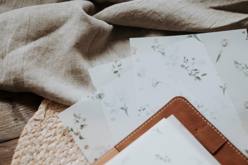 Puede incluir: Un diario de cuero marr&oacute;n con un inserto de papel blanco. El diario est&aacute; sobre una pila de hojas de papel blanco con un dise&ntilde;o floral. Las hojas est&aacute;n sobre una estera de paja tejida.