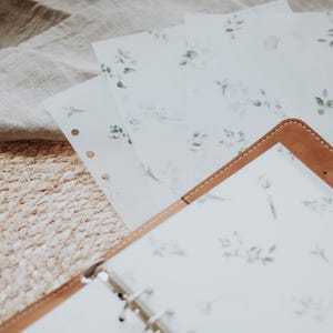 Puede incluir: Un diario encuadernado en cuero marr&oacute;n con una p&aacute;gina blanca con un delicado patr&oacute;n floral. El diario est&aacute; abierto y sobre una estera de paja tejida con varias hojas de papel con el mismo patr&oacute;n floral en la parte superior.