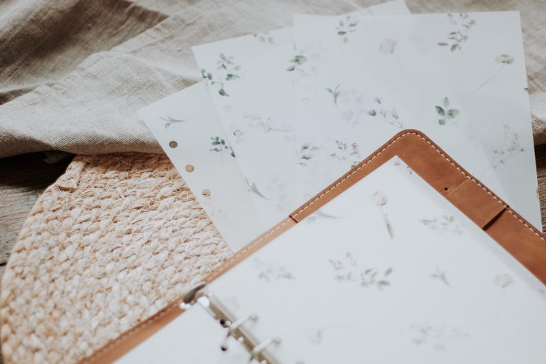 Puede incluir: Un diario encuadernado en cuero marr&oacute;n con una p&aacute;gina blanca con un delicado patr&oacute;n floral. El diario est&aacute; abierto y sobre una estera de paja tejida con varias hojas de papel con el mismo patr&oacute;n floral en la parte superior.