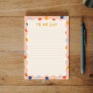 May include: A rectangular notepad with a scalloped edge and a light pink border featuring colorful polka dots. The center is cream-colored with horizontal lines and the text TO DO LIST printed in yellow at the top, accompanied by a gray pen.
