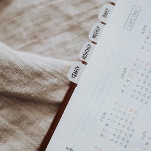 Peut inclure: Agenda ouvert avec des onglets indiquant "Annuel", "Mensuel", "Hebdomadaire", "Quotidien" et "Notes". Les pages du calendrier affichent l'année 2024, avec les mois de janvier, février et mars visibles. L'agenda est posé sur une surface claire.
