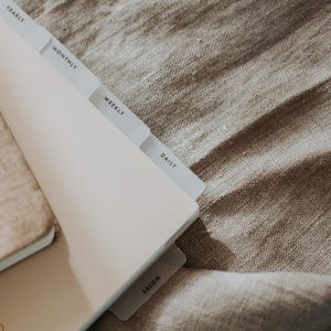 Op de afbeelding: Een lichtgekleurde planner met gelabelde tabbladen voor jaarlijkse, maandelijkse, wekelijkse, dagelijkse en notitiesecties. De planner is open en onthult de tabbladen tegen een neutraal getinte stoffen achtergrond.