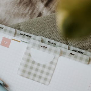 Pode incluir: Uma agenda com papel quadriculado branco e separadores com padrão vichy verde claro. Os separadores estão marcados com os meses August, September, October, April, November, May, December e June em texto preto. Uma caneta azul clara sobre a página.