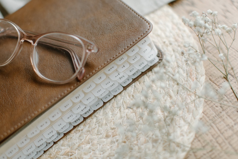 May include: A brown leather-bound journal with white tabs labelled with numbers and letters. The journal is lying on a woven mat with white flowers.