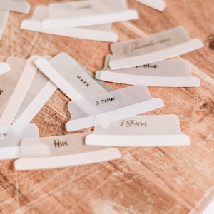 Op de afbeelding: Een close-up van een houten oppervlak met verschillende doorzichtige plastic scheiders met verschillende boektitels, waaronder "Markus", "1 Petrus", "3 Johannes", "Titus" en "2 Thessalonicenzen".