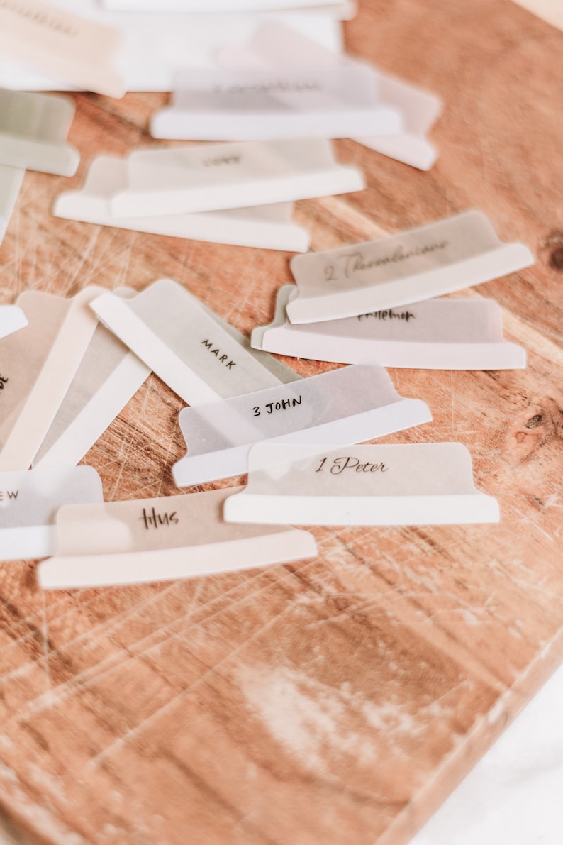 Op de afbeelding: Een close-up van een houten oppervlak met verschillende doorzichtige plastic scheiders met verschillende boektitels, waaronder "Markus", "1 Petrus", "3 Johannes", "Titus" en "2 Thessalonicenzen".