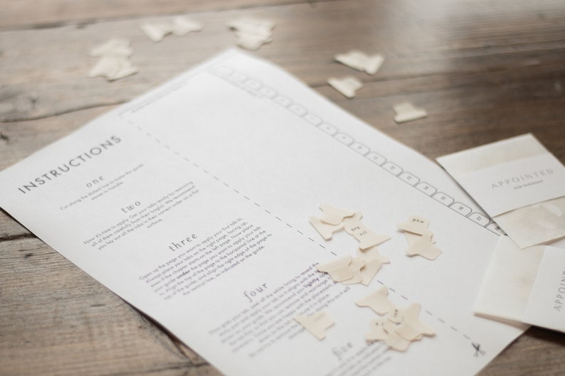 May include: A white sheet of paper with instructions for a game called "Appointed". The instructions are numbered one to five. There are small white paper shapes with numbers on them scattered on the paper. The paper is on a wooden surface.