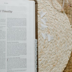 May include: A close-up of an open Bible with a brown leather cover and white page tabs. The Bible is open to the book of 1 Timothy, chapter 1, verses 1-2. The text is visible and includes the words "trustworthy and appointed me to serve him, even though I used to blaspheme the name of Christ." The Bible is resting on a woven straw mat with a pair of clear glasses resting on top.