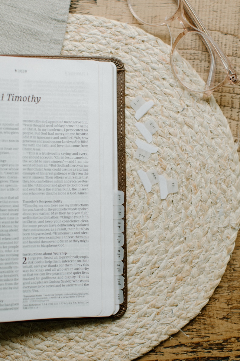 May include: A close-up of an open Bible with a brown leather cover and white page tabs. The Bible is open to the book of 1 Timothy, chapter 1, verses 1-2. The text is visible and includes the words "trustworthy and appointed me to serve him, even though I used to blaspheme the name of Christ." The Bible is resting on a woven straw mat with a pair of clear glasses resting on top.