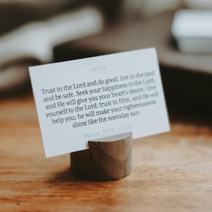 May include: A white card with text from Psalm 37:3-6, held in a small, wooden holder. The card's text is in black, and the holder is a natural wood colour. The background is a blurred wooden surface.