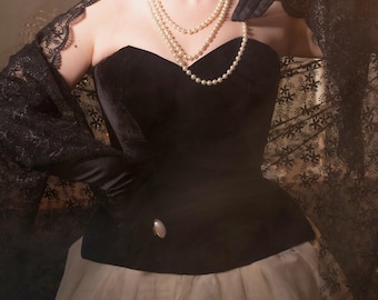 May include: A woman wearing a black velvet corset with a white tulle skirt and a black lace shawl. She is wearing a pearl necklace and earrings. The background is a soft brown with a spotlight shining on her.