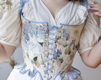 May include: A floral corset featuring a light blue and white pattern with a front lace-up closure using blue cord. The corset has a scalloped hem with blue trim and is worn over a white blouse with puffed sleeves and a gathered skirt.