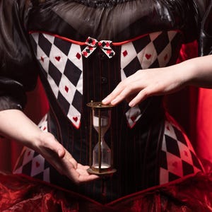 May include: A black and white corset with a diamond and heart pattern, red trim, and a small bow. The corset is paired with a sheer black top and red skirt. A brass hourglass is held in front of the corset.