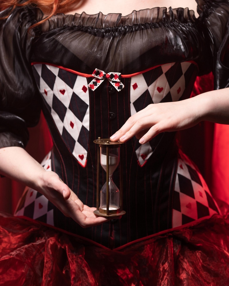 May include: A black and white corset with a diamond and heart pattern, red trim, and a small bow. The corset is paired with a sheer black top and red skirt. A brass hourglass is held in front of the corset.