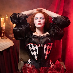 May include: A black corset with a harlequin pattern of black and white diamonds and red hearts. The corset is paired with a black top with puffy sleeves and a red skirt. The background is a dark red with candles.