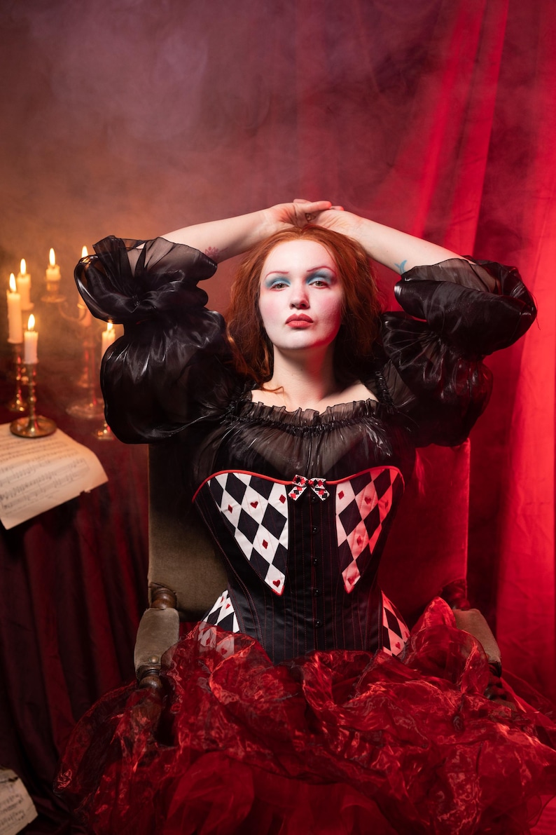 May include: A black corset with a harlequin pattern of black and white diamonds and red hearts. The corset is paired with a black top with puffy sleeves and a red skirt. The background is a dark red with candles.