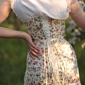 May include: A floral corset with red and pink roses on a cream background. The corset has a lace-up back and is paired with a white top and skirt. The image is taken outdoors in natural light.