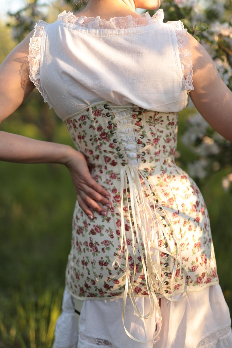 May include: A floral corset with red and pink roses on a cream background. The corset has a lace-up back and is paired with a white top and skirt. The image is taken outdoors in natural light.
