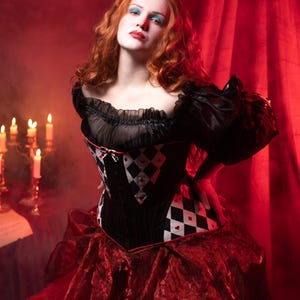 May include: A woman in a black and red costume with a corset featuring a diamond pattern. The costume has a ruffled skirt and sleeves. The background is a red curtain and candles.
