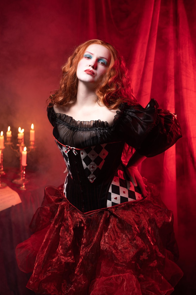 May include: A woman in a black and red costume with a corset featuring a diamond pattern. The costume has a ruffled skirt and sleeves. The background is a red curtain and candles.