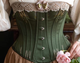 May include: An olive green corset with a decorative lace trim and a small bow. The corset has silver-colored clasps down the front and is paired with a tan skirt. A pink rose is held in the hand.