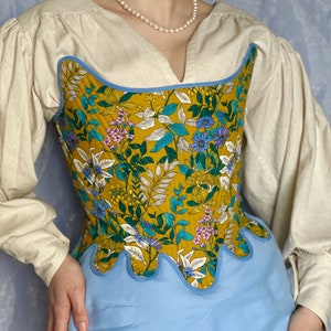 Peut inclure: Un corset floral jaune avec une bordure bleue et un bas festonné. Le corset est porté sur une blouse blanche.