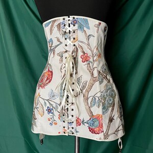 May include: A cream-colored corset with a floral and bird pattern. The corset features a lace-up front with cream-colored ribbon and metal eyelets. The design includes red, blue, and orange flowers and birds on branches. The corset has garter straps.