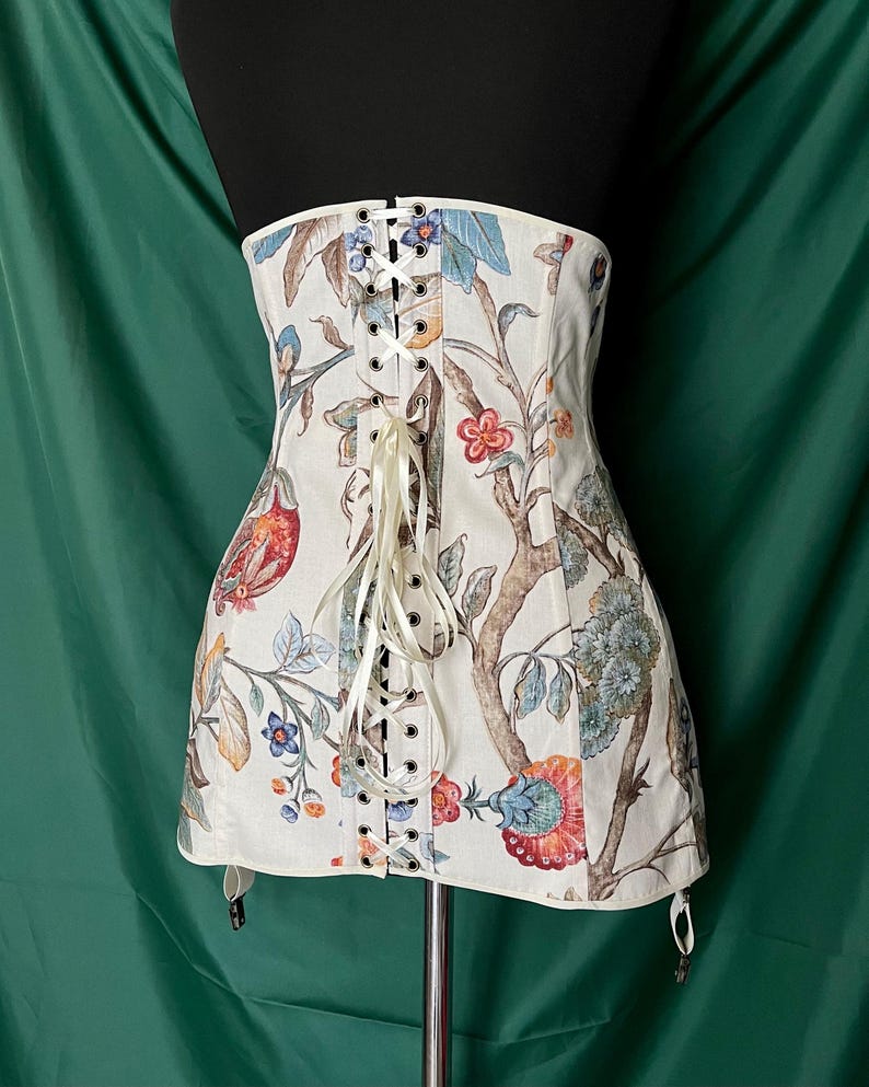 May include: A cream-colored corset with a floral and bird pattern. The corset features a lace-up front with cream-colored ribbon and metal eyelets. The design includes red, blue, and orange flowers and birds on branches. The corset has garter straps.