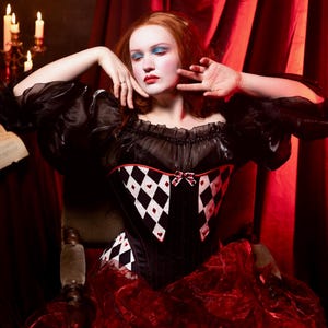 May include: A black corset with a harlequin pattern of white diamonds and red hearts. The corset has a sweetheart neckline and is paired with a red ruffled skirt. The model is wearing a black off-the-shoulder top with puffy sleeves.