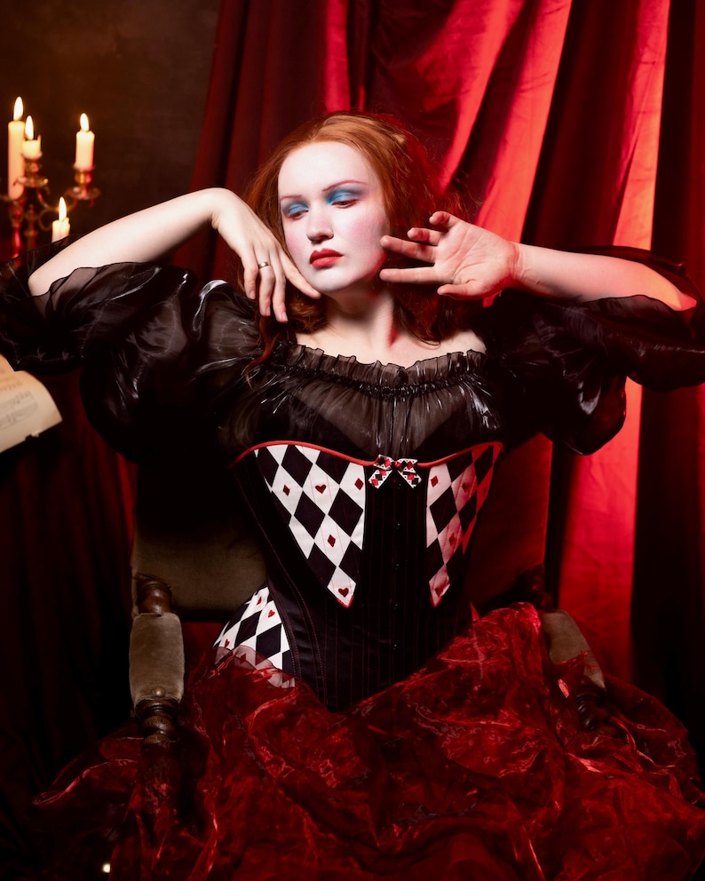 May include: A black corset with a harlequin pattern of white diamonds and red hearts. The corset has a sweetheart neckline and is paired with a red ruffled skirt. The model is wearing a black off-the-shoulder top with puffy sleeves.
