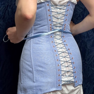May include: A light blue corset with a floral pattern and copper-colored eyelets. The corset is laced up the back with blue ribbon. The garment is worn over a white top and skirt. The overall style is vintage.